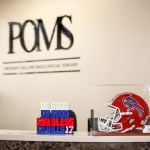 Modern dental reception area with white counter, “POMS” logo on wall, potted plants, and wood-look flooring